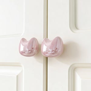 May include: Two pink, cat-shaped cabinet knobs with a glossy finish are mounted on a white cabinet door. The knobs have a rounded, smooth shape with a subtle iridescent sheen, reflecting light. The cabinet door has a recessed panel design.