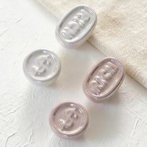 May include: Four ceramic knobs in shades of white and pink. Two oval knobs read "RICH" in raised letters. The other two are round with a horse design. The knobs are likely for furniture or decor.