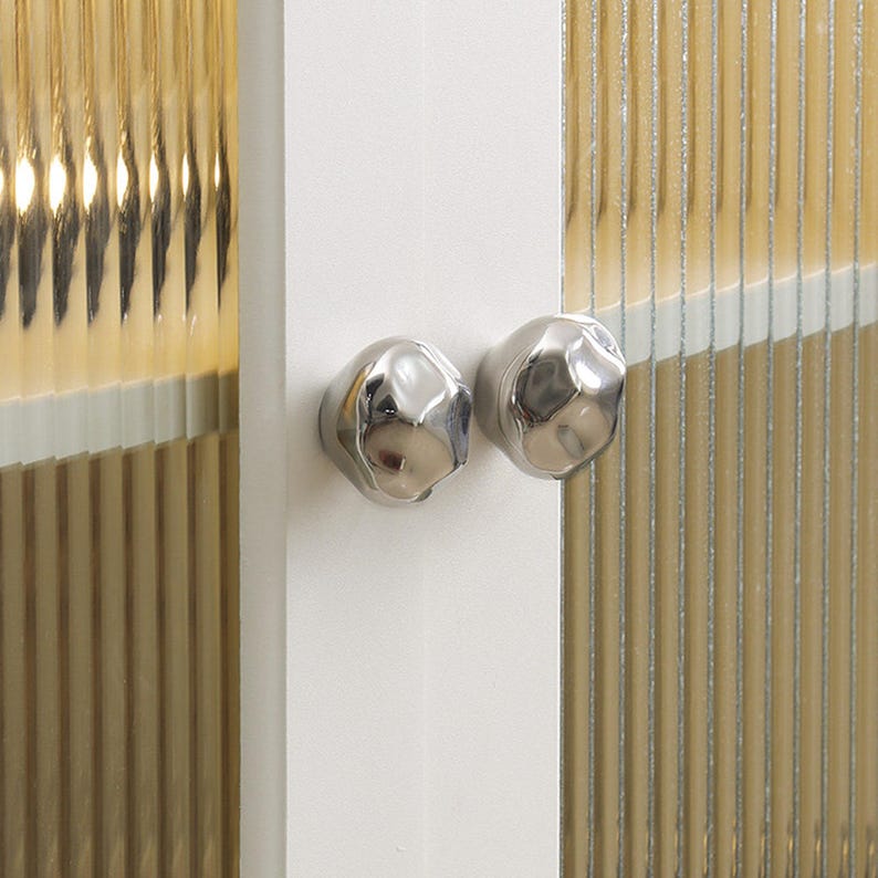 May include: A white cabinet door with two silver, organic-shaped knobs. Adjacent is a panel of ribbed, translucent glass with a warm, golden colour. The knobs have a reflective, metallic finish. The glass panel is made of vertical lines.