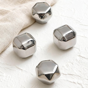 May include: Four silver-coloured, faceted decorative objects. Each object has a geometric, gem-like shape with multiple flat surfaces that reflect light. The objects are arranged on a white surface with a neutral-coloured cloth in the background.