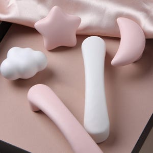 以下が含まれることがあります： 星、雲、三日月、2つの細長い取っ手など、さまざまな形のパステルカラーのキャビネットノブのコレクション。 ノブはソフトピンクと白で、ピンクの表面に配置されています。