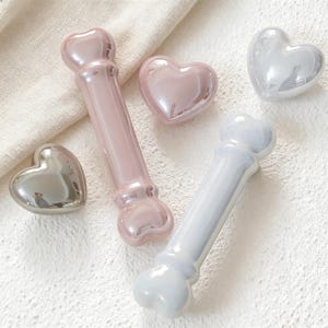 Puede incluir: Una colección de piezas decorativas de cerámica con forma de corazón y hueso. Las piezas son en tonos rosa, plata y blanco, con un acabado brillante. La disposición incluye dos piezas en forma de hueso y tres piezas en forma de corazón.