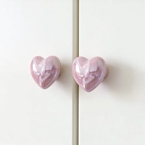 May include: Two pink heart-shaped cabinet knobs with a glossy finish are mounted on a white cabinet door. The knobs are symmetrical and reflect light, adding a touch of charm to the furniture. The knobs are approximately 2 inches in diameter.