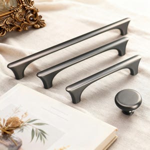 Peut inclure: Un ensemble de quatre pièces de quincaillerie d'armoire gris foncé. L'ensemble comprend trois poignées de tirage de différentes tailles et un bouton rond. Le matériel est en métal et présente un design moderne et minimaliste. Les pièces sont disposées sur une surface de couleur neutre.