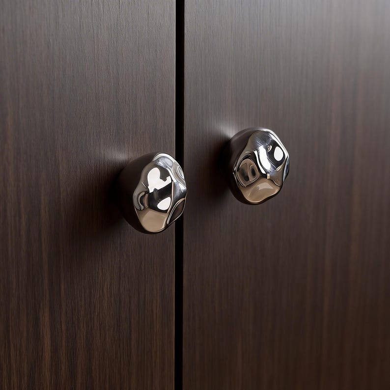 May include: Two polished chrome cabinet knobs with an organic, textured design are mounted on a dark brown wooden cabinet door. The knobs reflect light, creating a contrast in colour and texture. The cabinet door is made of wood.
