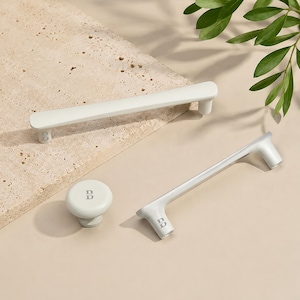 May include: Two matte off-white cabinet pulls and one matching round cabinet knob. Each piece features a small, embossed letter B on the base. The hardware has a smooth, elongated design with rounded edges and a clean, uniform finish throughout.