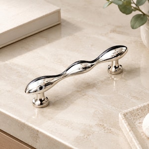 May include: A polished silver-tone metal cabinet handle featuring a smooth, undulating wave-like design. The handle has a reflective, mirror-like finish and is supported by two cylindrical bases that connect to the main body at each end.