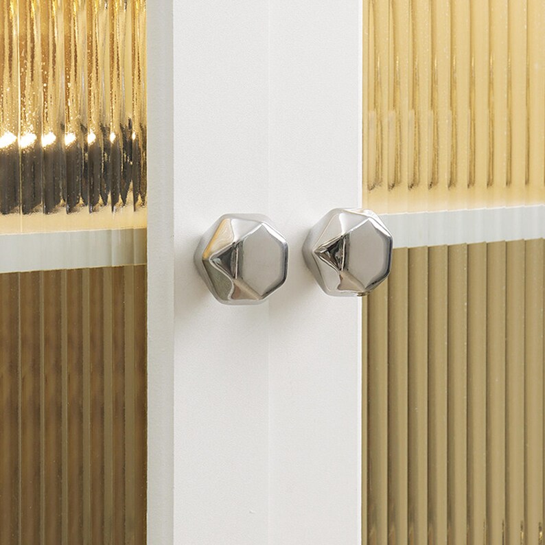 Op de afbeelding: Close-up van twee zilveren, geometrische kastknoppen gemonteerd op een witte kastdeur. De achtergrond toont een getextureerd, goudkleurig glazen paneel. De knoppen hebben een facetontwerp dat licht reflecteert.