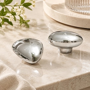 May include: Two polished silver-toned cabinet knobs with a smooth, rounded, pebble-like shape. Each knob features a subtle, etched letter A centered on the reflective surface. The hardware has a metallic finish that creates a mirror-like effect.