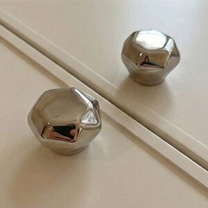 Op de afbeelding: Twee gepolijste zilveren kastknoppen met een facet, geometrisch ontwerp. De knoppen zijn gemonteerd op een witte kastdeur, die licht reflecteren en een moderne touch aan het meubel geven.