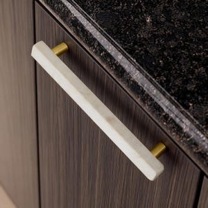 May include: A white marble cabinet pull with brass-coloured accents. The rectangular pull is mounted on a dark wood cabinet with a black countertop. The hardware is a modern design element.