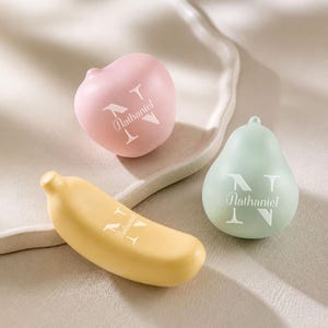 Könnte beinhalten: Drei glatte, mattierte Objekte in Fruchtform: ein rosa Apfel, eine hellgrüne Birne und eine gelbe Banane. Jeder Gegenstand zeigt ein weißes Druckdesign, bestehend aus einem großen Buchstaben N mit dem Namen Nathaniel in Schreibschrift.