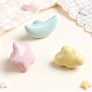 May include: Three ceramic cabinet knobs shaped like a crescent moon, star, and cloud. The moon is light blue, the star is pink, and the cloud is yellow. These decorative knobs are suitable for a nursery or children's room.