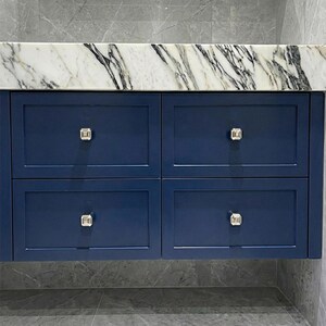 Op de afbeelding: Een marineblauwe badkamermeubel met vier lades. De lades hebben zilveren vierkante knoppen en zijn afgewerkt met een wit en grijs marmeren aanrechtblad. De meubel is gemonteerd op een grijze betegelde muur.
