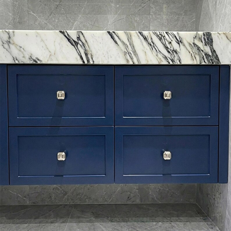 Op de afbeelding: Een marineblauwe badkamermeubel met vier lades. De lades hebben zilveren vierkante knoppen en zijn afgewerkt met een wit en grijs marmeren aanrechtblad. De meubel is gemonteerd op een grijze betegelde muur.