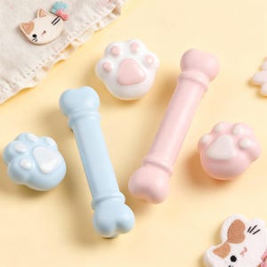 May include: A selection of pet-themed accessories, featuring two bone-shaped toys in light blue and pink, and paw-shaped items in white and pink. A cat-shaped embellishment is also visible. The items are arranged on a light yellow surface.
