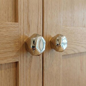 Op de afbeelding: Close-up van twee goudkleurige, gefacetteerde kastknoppen gemonteerd op een lichtgekleurde houten kastdeur. De knoppen hebben een reflecterend oppervlak en de houtnerf is zichtbaar.