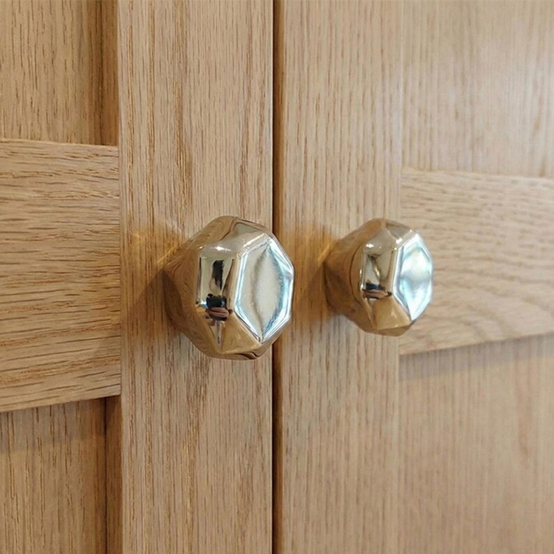 Op de afbeelding: Close-up van twee goudkleurige, gefacetteerde kastknoppen gemonteerd op een lichtgekleurde houten kastdeur. De knoppen hebben een reflecterend oppervlak en de houtnerf is zichtbaar.