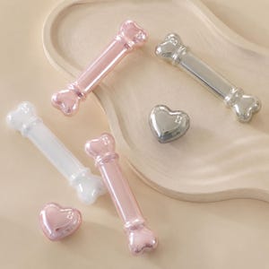 May include: A collection of small, smooth, metallic-finish objects including three bone-shaped pieces in metallic pink, silver, and white, and two heart-shaped pieces in metallic pink and silver, each featuring a small letter B embossed on the surface.