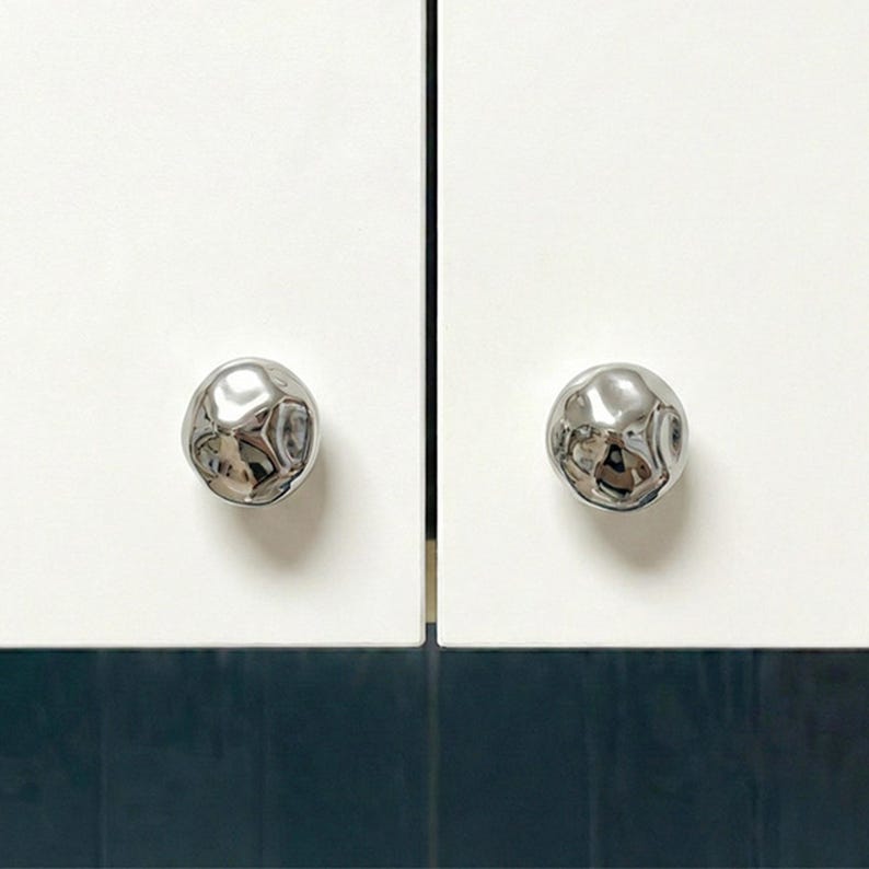 May include: Two white cabinet doors with shiny, textured, silver-coloured knobs. The knobs have an organic, irregular shape. The cabinet doors are above a dark blue surface, creating a modern, minimalist aesthetic.