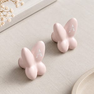 May include: Two light pink cabinet knobs shaped like butterflies. Each knob features a smooth, rounded surface with a white letter A printed on the upper wing. The knobs have a solid, matte finish and a three-dimensional form with distinct wing sections.