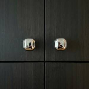 Op de afbeelding: Close-up van twee glanzende, zilveren kastknoppen met een achthoekige vorm. De knoppen zijn gemonteerd op een donkere kastdeur met houtnerf. De knoppen reflecteren de omgeving.