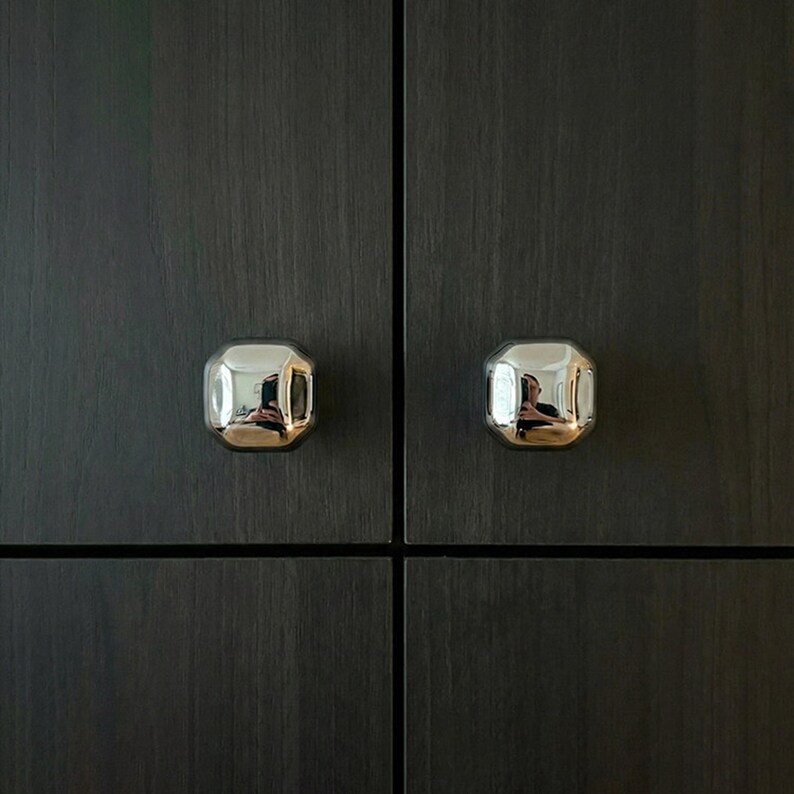 Op de afbeelding: Close-up van twee glanzende, zilveren kastknoppen met een achthoekige vorm. De knoppen zijn gemonteerd op een donkere kastdeur met houtnerf. De knoppen reflecteren de omgeving.