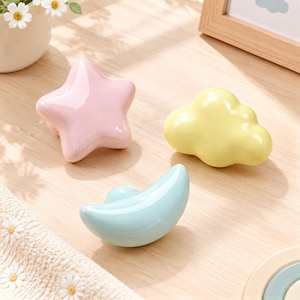 Puede incluir: Tres pomos de cerámica para cajones en colores pastel: una estrella rosa claro, una nube amarilla pálida y una luna creciente azul claro. Cada pieza tiene un acabado liso y brillante con una forma redondeada para muebles.