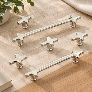 Puede incluir: Un conjunto de herrajes de metal plateado pulido que incluye dos tiradores largos para cajones y tres pomos individuales. Cada pieza presenta un diseño en forma de cruz con cuatro brazos redondeados que parten de una base cilíndrica central.