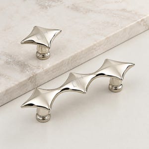 May include: A polished silver-toned metal cabinet knob and a matching elongated cabinet pull handle. Both pieces feature a distinct four-pointed star shape with a smooth, reflective surface and a cylindrical base for mounting to furniture or cabinetry.