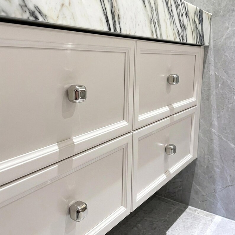 Op de afbeelding: Close-up van een witte badkamermeubel met vier lades. Elke lade heeft een zilveren, vierkante knop. De wastafel staat onder een marmeren aanrecht met grijze en witte aderen. De muur is grijs.