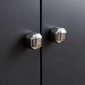 Op de afbeelding: Twee gepolijste chromen kastknoppen met een geometrisch ontwerp zijn gemonteerd op een donkergrijze kastdeur. De knoppen hebben een reflecterend oppervlak en de deur is effen van kleur.