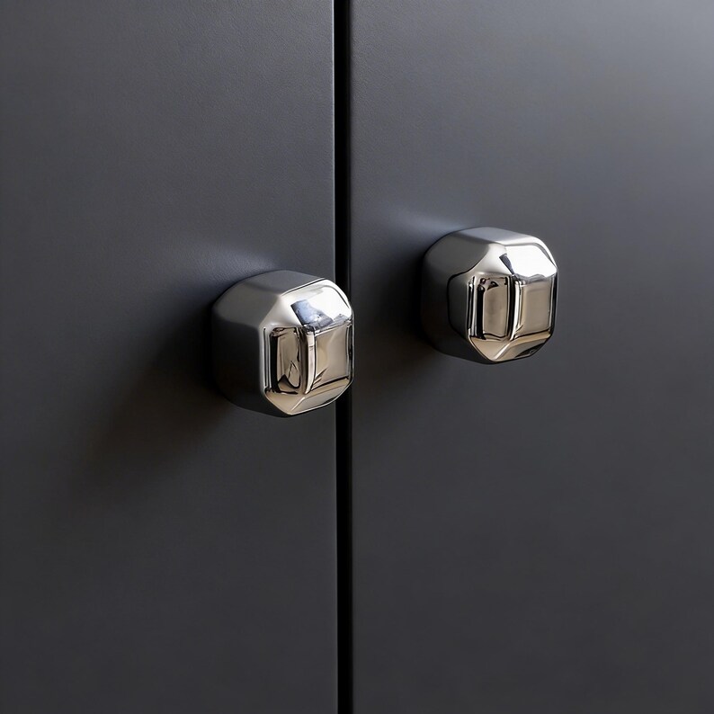 Op de afbeelding: Twee gepolijste chromen kastknoppen met een geometrisch ontwerp zijn gemonteerd op een donkergrijze kastdeur. De knoppen hebben een reflecterend oppervlak en de deur is effen van kleur.
