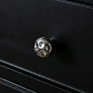 May include: A close-up of a silver, textured cabinet knob with a hammered design. The knob is round and reflects light, set against a black wooden surface with horizontal grooves. Furniture hardware.