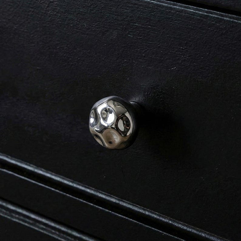 May include: A close-up of a silver, textured cabinet knob with a hammered design. The knob is round and reflects light, set against a black wooden surface with horizontal grooves. Furniture hardware.