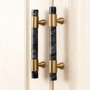 May include: Two cabinet pulls with a cylindrical design. The pulls feature a dark gray marble center section, accented by gold-colored metal rings and curved end pieces. The pulls are mounted on a white cabinet door.
