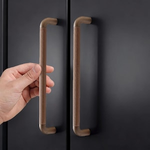 May include: Close-up of two brown cabinet handles with a textured grip. The handles are mounted on a dark gray cabinet door. One hand is reaching for a handle.