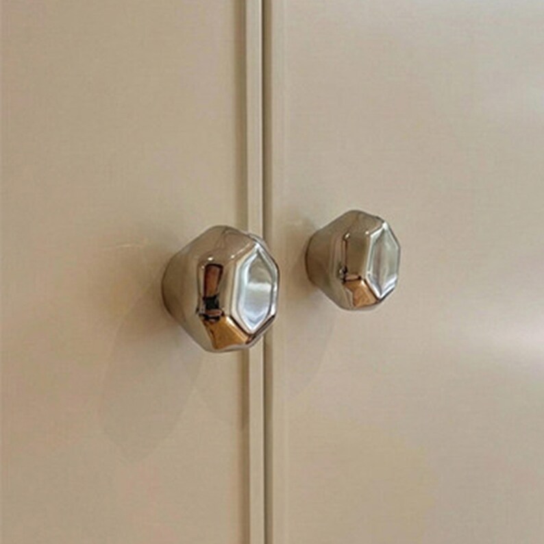 Op de afbeelding: Twee gepolijste chromen kastknoppen met een facetvormige, organische vorm. De knoppen zijn gemonteerd op een lichtbeige kastdeur, die de omgeving weerspiegelt. De hardware heeft een moderne, minimalistische uitstraling.