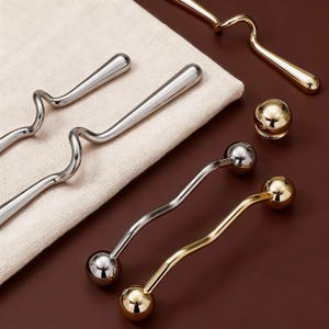 Puede incluir: Una variedad de herramientas de bar, que incluyen agitadores plateados y dorados con diseños curvos y esféricos. También hay un pequeño pomo esférico dorado. Las herramientas están dispuestas sobre un paño de color neutro y una superficie marrón.