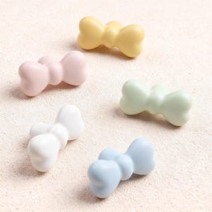May include: Five pastel-coloured bow-shaped objects. The bows are in shades of yellow, pink, white, green, and blue. The objects are smooth and appear to be made of a solid material.