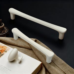 May include: Two off-white cabinet pulls and a matching knob are displayed against a dark background. The pulls are long and rectangular with rounded ends. The knob is oval-shaped with a short, cylindrical base. The hardware is likely for kitchen or bathroom cabinets.