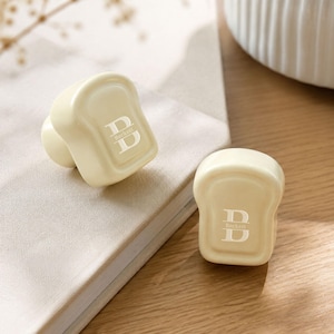 May include: Two cream-coloured, bread-shaped knobs featuring a white printed design of a large letter B with the name Beckett written horizontally across the centre. The knobs have a smooth, rounded finish and a raised border around the front face.