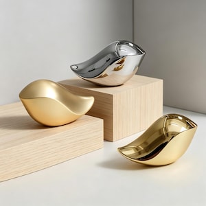 May include: Three bird-shaped decorative figurines with smooth, reflective surfaces. One figurine has a matte gold finish, one has a polished silver finish, and one has a polished gold finish. Each bird is stylized with a rounded body and a pointed tail.