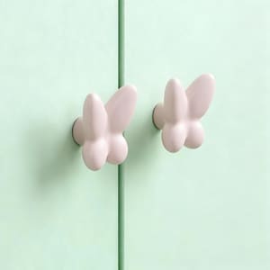 以下が含まれることがあります： 淡いピンク色の蝶の形をした2つのキャビネットノブが、明るい緑色のキャビネットドアに取り付けられています。ノブは丸みを帯びた3次元のデザインです。2つのノブの間には、垂直の緑色の線があります。
