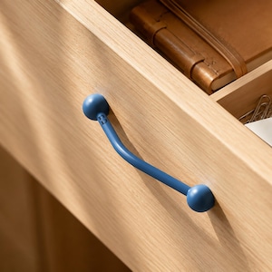 May include: A matte blue metal drawer pull with a curved bar design and spherical ends, mounted horizontally on a light-toned wooden drawer front. The handle features a smooth finish and a simple, clean aesthetic, contrasting with the natural wood grain.