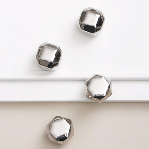 Op de afbeelding: Vier glanzende, zilveren kastknoppen met een facet, geometrisch ontwerp zijn gerangschikt op een witte en cr&egrave;mekleurige ondergrond. De knoppen hebben een reflecterende, metalen afwerking.