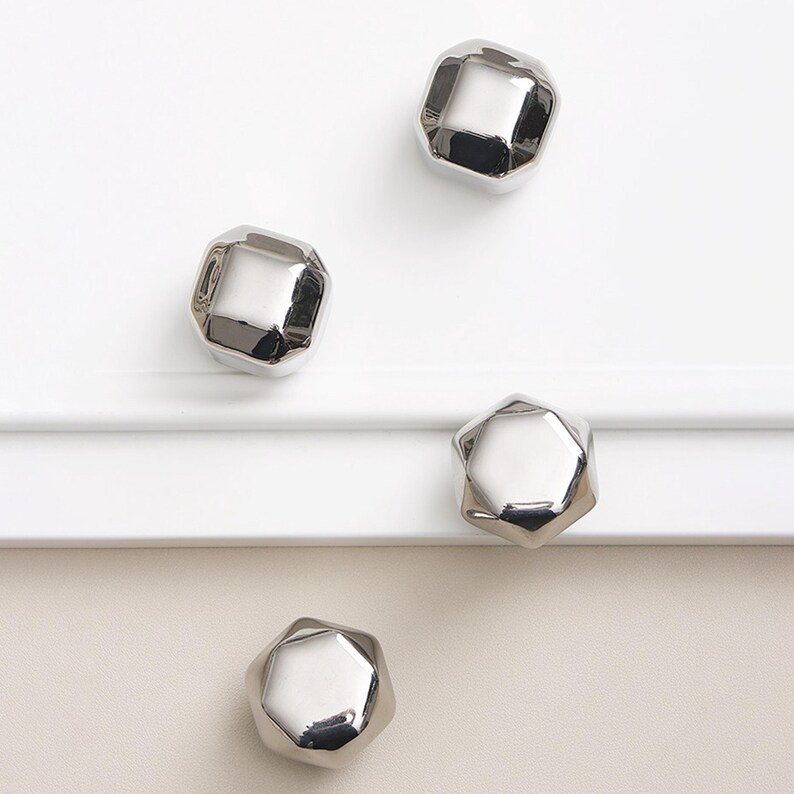 Op de afbeelding: Vier glanzende, zilveren kastknoppen met een facet, geometrisch ontwerp zijn gerangschikt op een witte en cr&egrave;mekleurige ondergrond. De knoppen hebben een reflecterende, metalen afwerking.