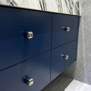 Op de afbeelding: Een close-up van een marineblauwe badkamermeubel met vier lades. Elke lade heeft een zilveren, achthoekige knop. De meubel heeft een wit en grijs marmeren aanrechtblad.