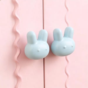 May include: Two pale blue bunny-shaped cabinet knobs on a pink cabinet door. The knobs have a smooth, rounded design with small details for eyes and a nose. The cabinet door is a soft pink colour.
