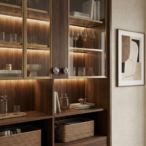 May include: A built-in wooden cabinet with glass doors and interior lighting. The cabinet displays glassware, books, and decorative items. The frame has a gold trim. A framed abstract art piece hangs on the wall to the right.